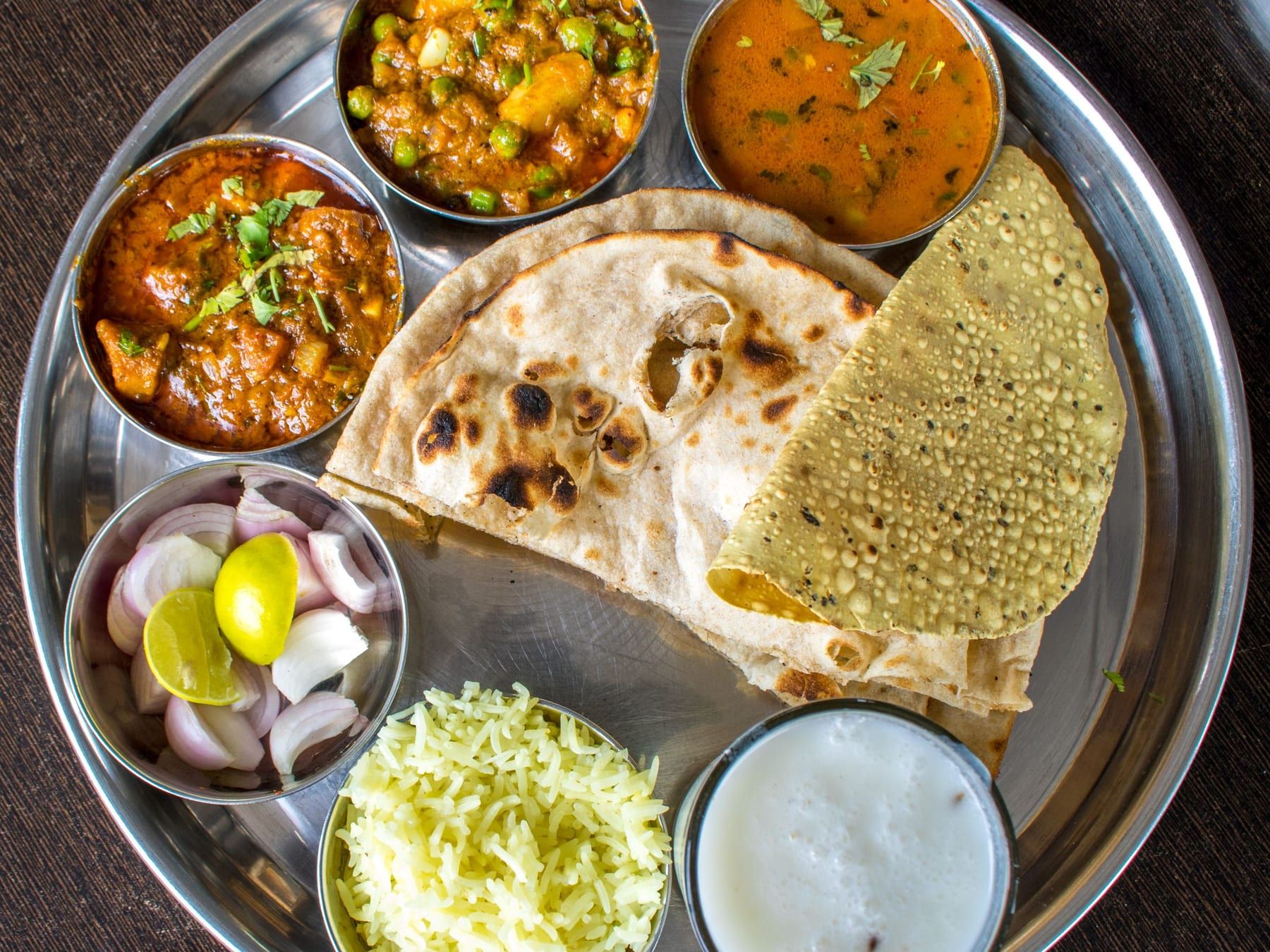 A steel thali with paneer curry, dal, rice, raita, roti, papad and sliced onion